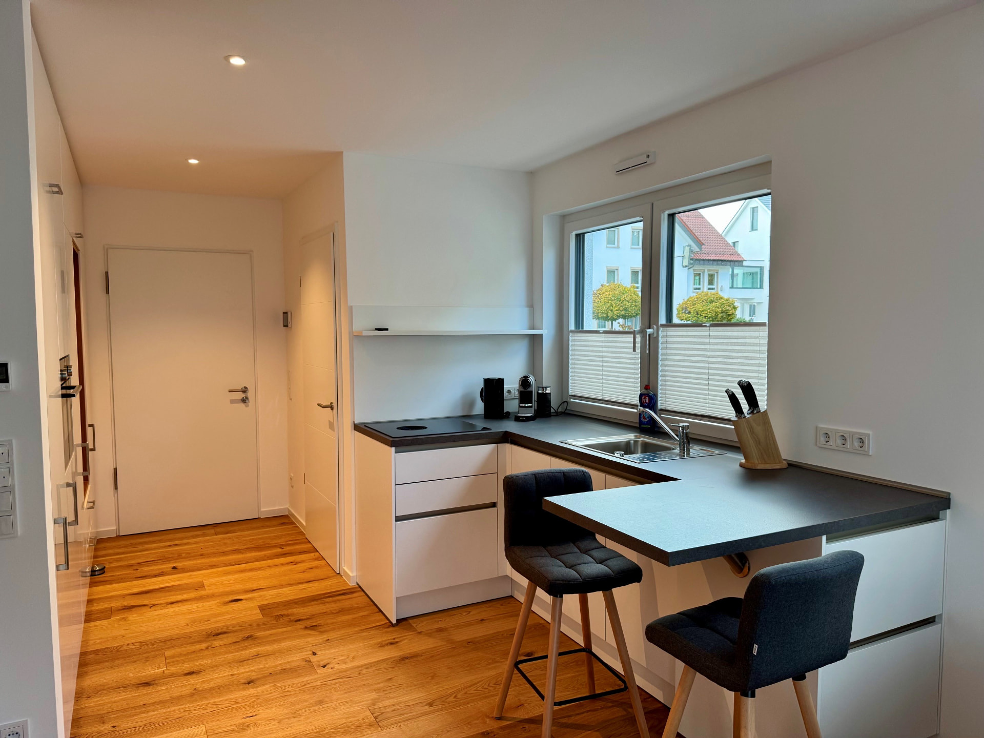 Küche mit weißer Front, dunkler Arbeitsplatte, Kochfeld, Spüle und Theke mit zwei Barhockern.