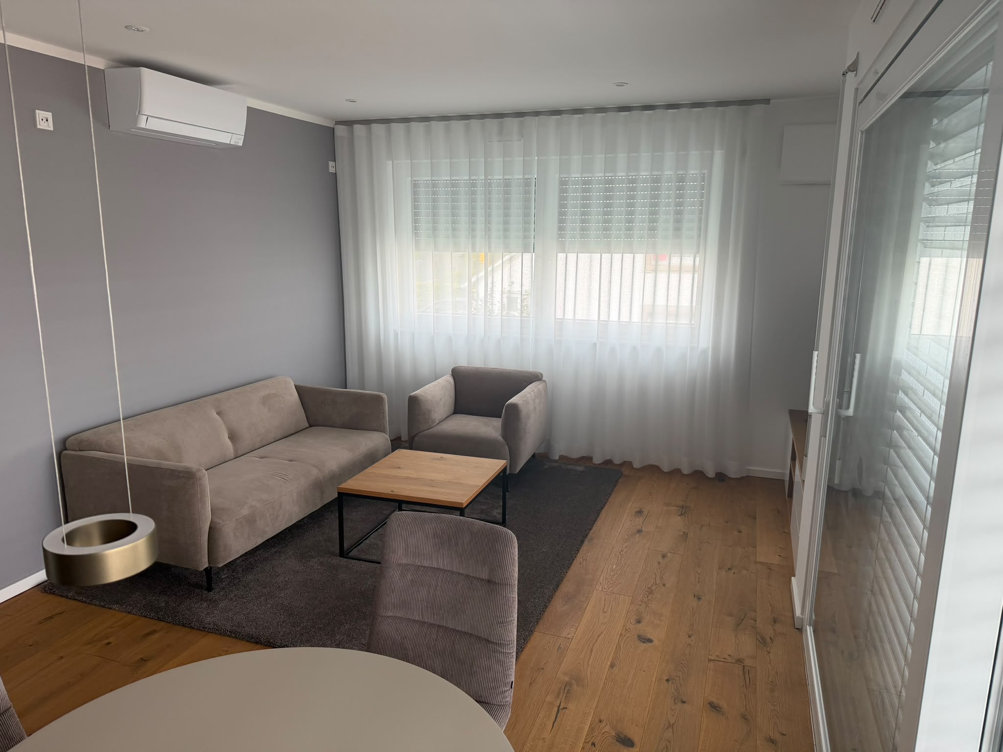 Wohnzimmer-Ecke mit beigem Sofa, Sessel, Couchtisch und bodentiefen Vorhängen vor der Fensterfront.