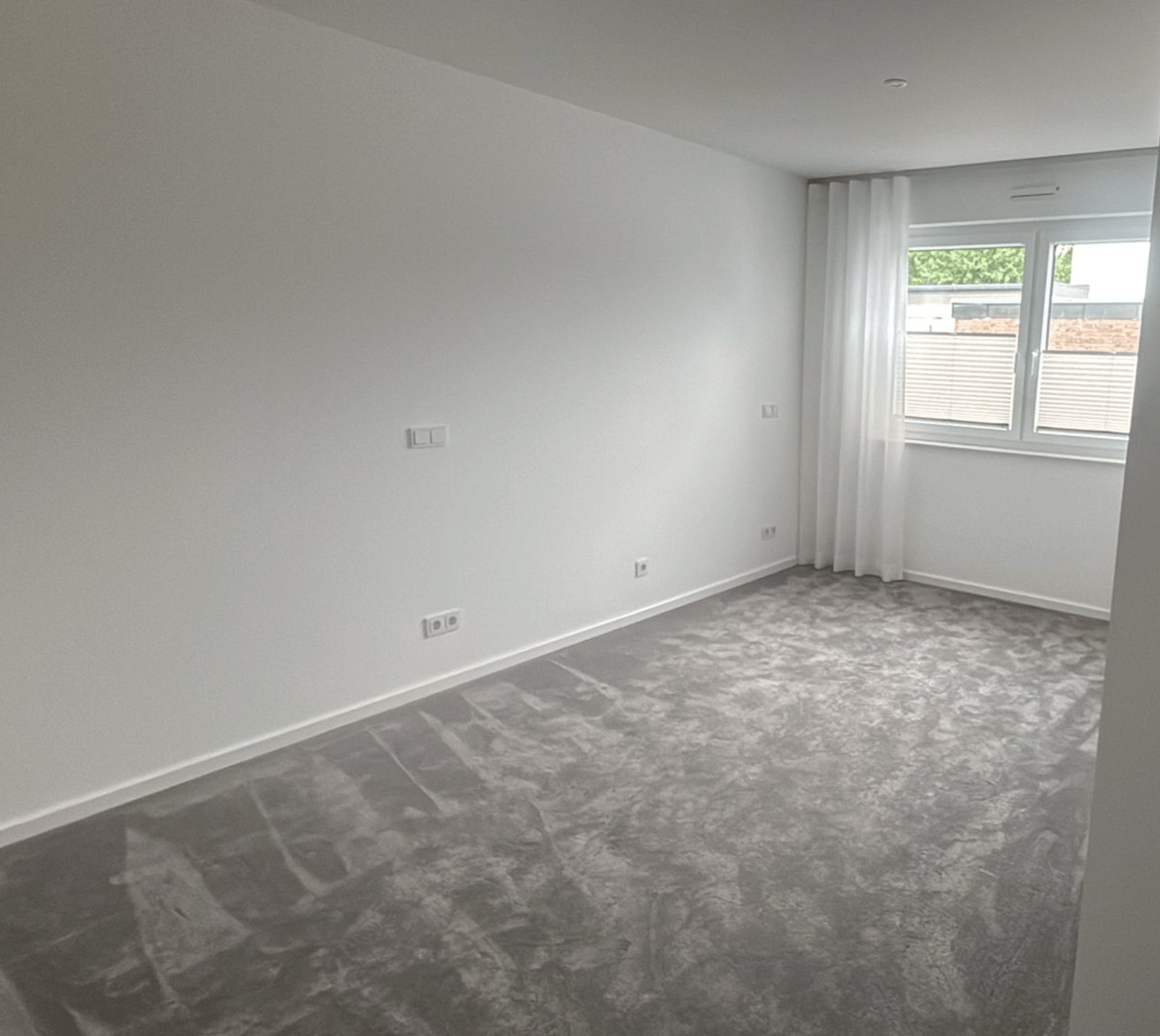 Leeres Zimmer mit weißen Wänden, grauem Teppichboden und einem Fenster mit weißen Vorhängen.