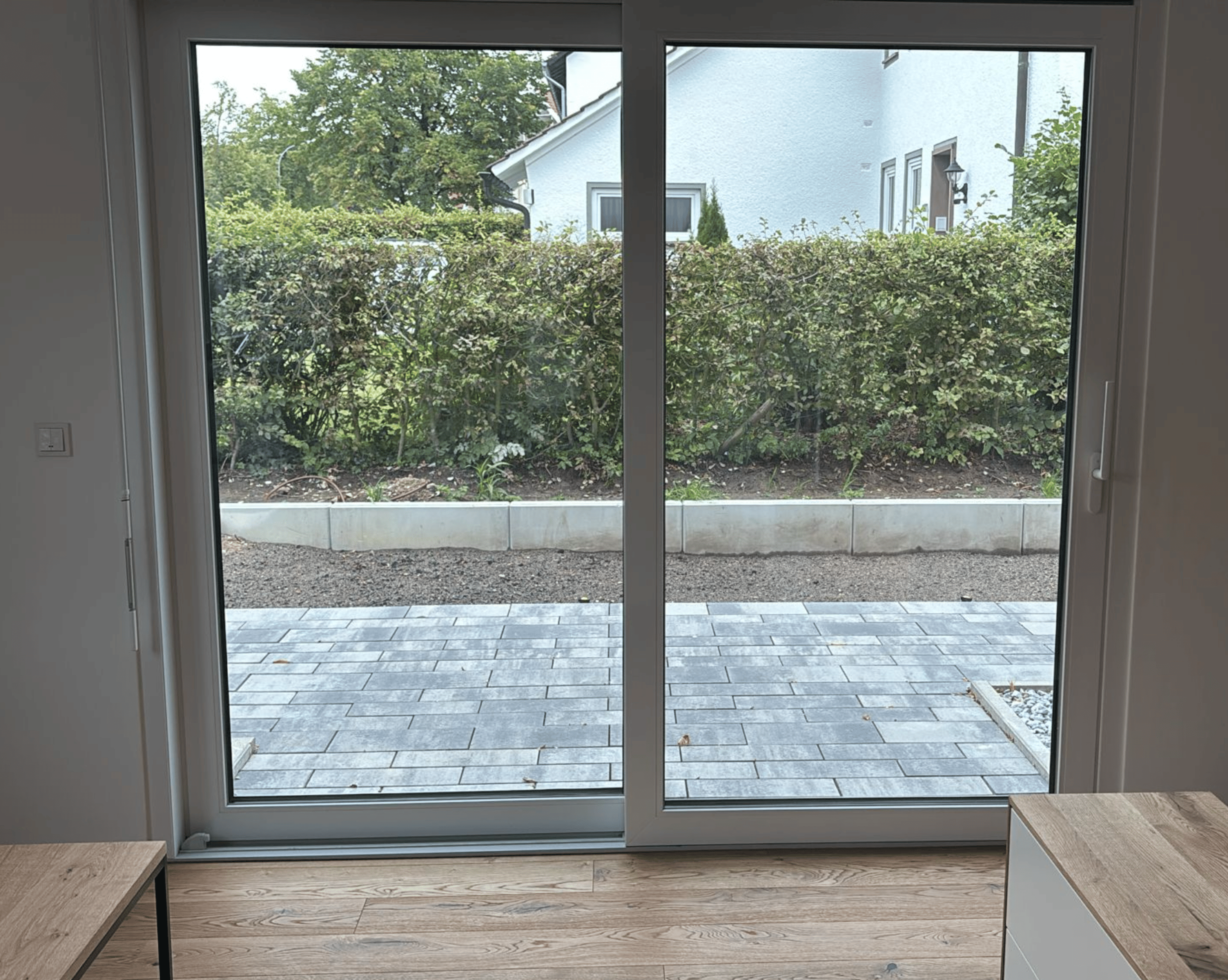 Blick durch eine Glasschiebetür auf eine gepflasterte Terrasse und eine Hecke vor einem Haus.