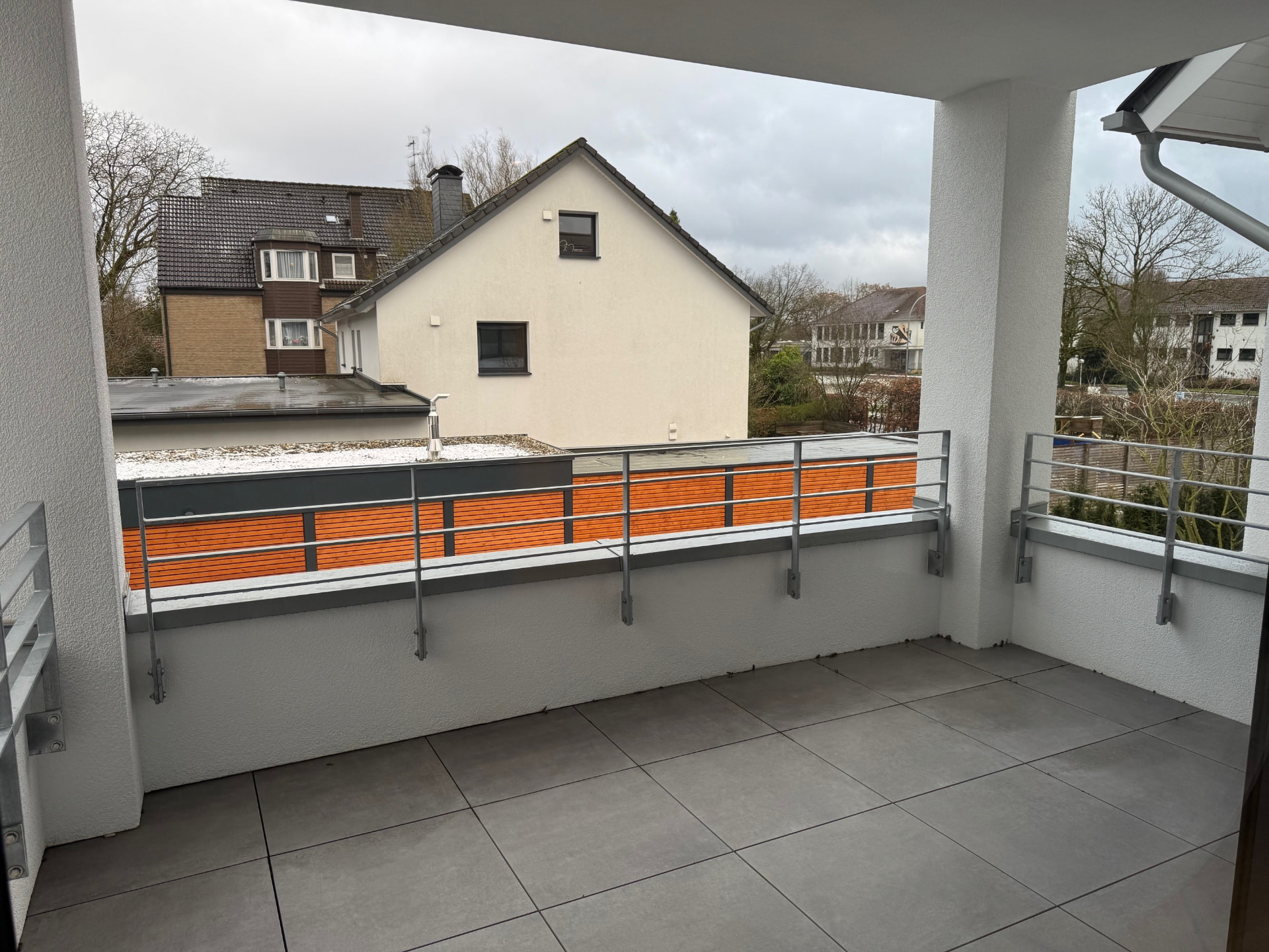 Überdachter Balkon mit grauen Bodenfliesen, Geländer und Blick ins Grüne.