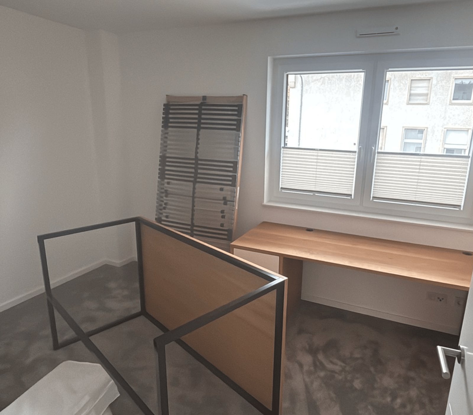 Leeres Zimmer mit grauem Teppich, einem Holztisch unter einem Fenster und einem Stuhl.
