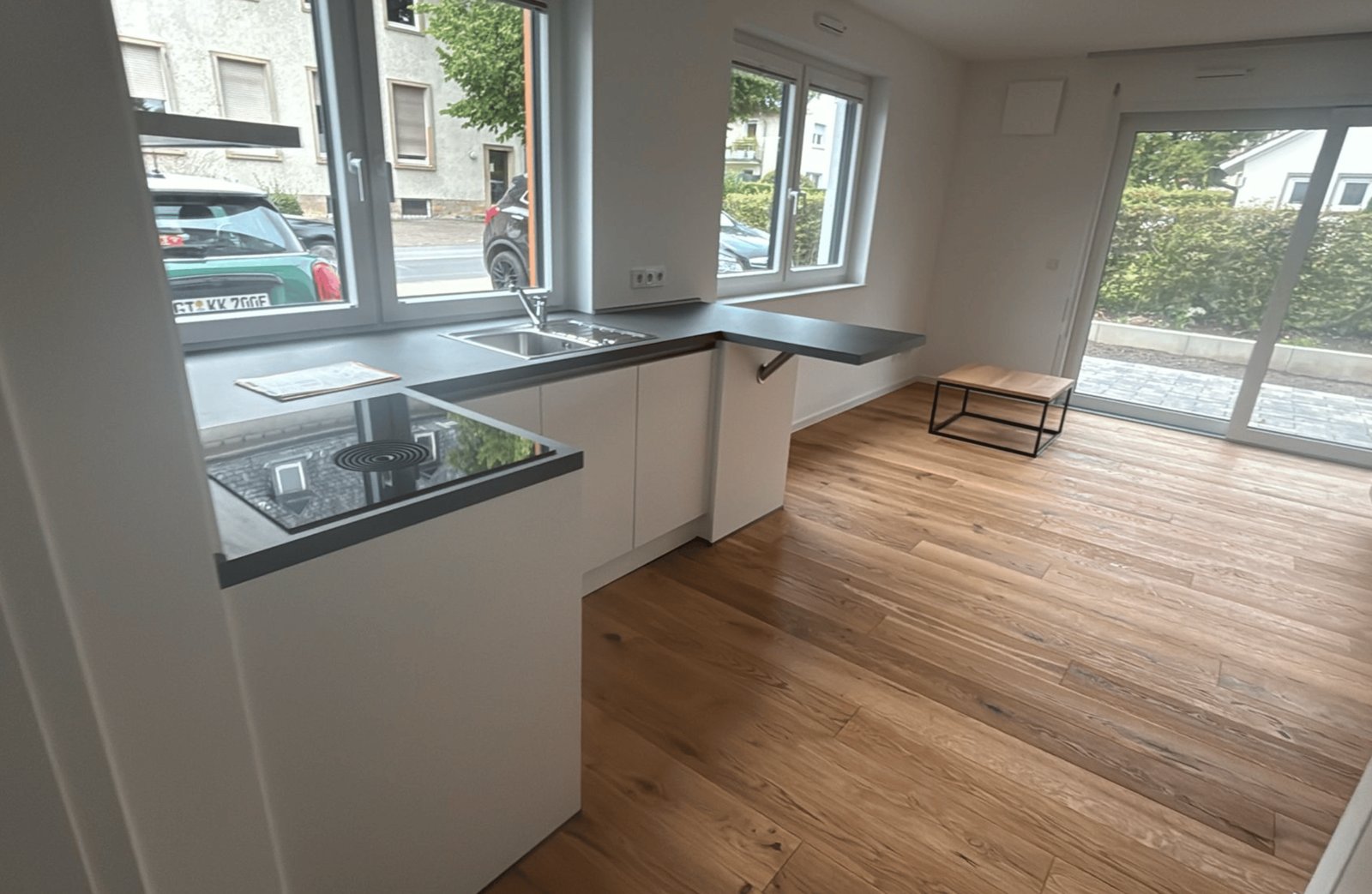 Eine moderne Wohnküche mit weißer Einbauküche, dunkler Arbeitsplatte, Holzboden und einem großen Fenster.