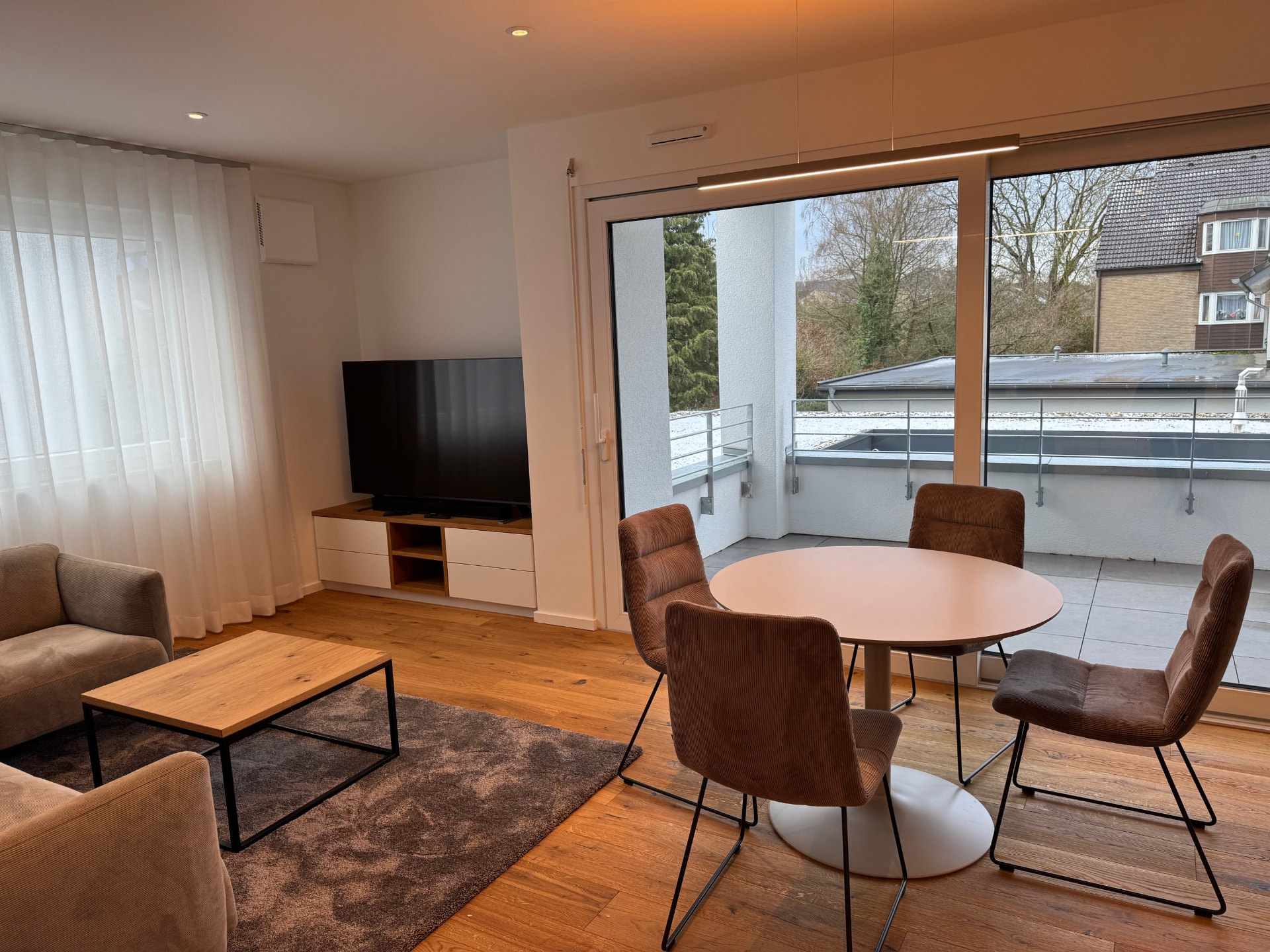 Wohnzimmer mit Sesseln, Couchtisch, TV-Sideboard und rundem Esstisch vor der Terrassentür.