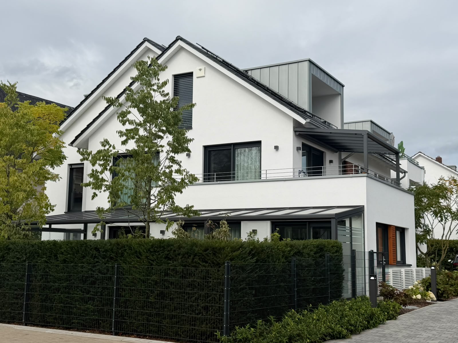 Gebäudeseite mit Dachterrasse, Metalldach-Pergola, Gaubenbalkon und Glasdach-Terrasse im Erdgeschoss.