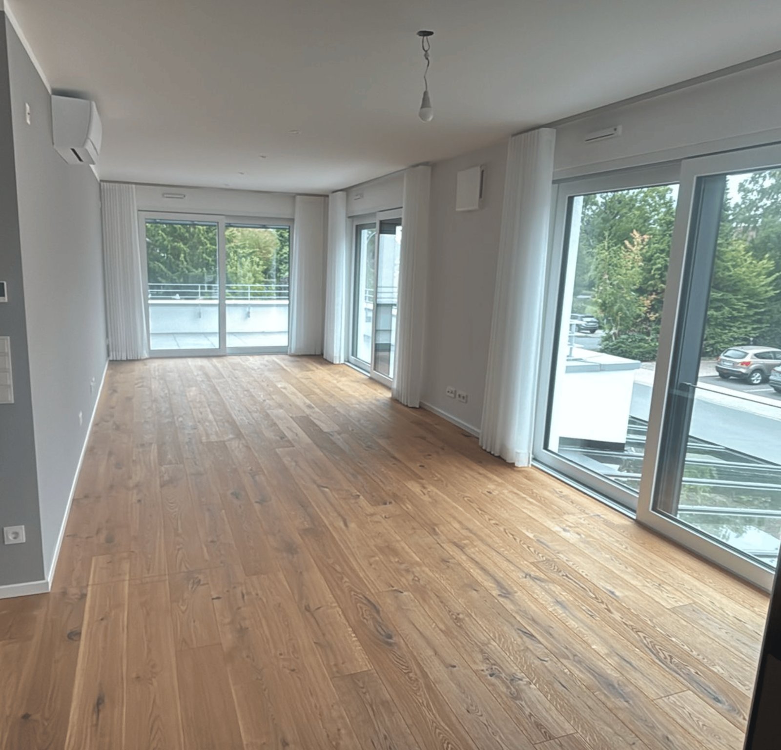 Leeres Zimmer mit Holzboden und großen Fenstern mit weißem Vorhang.