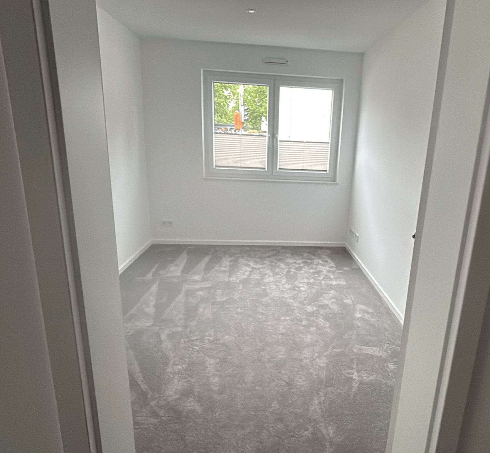 Leerer Raum mit weiß gestrichenen Wänden, grauem Teppichboden und einem Fenster in der Mitte.