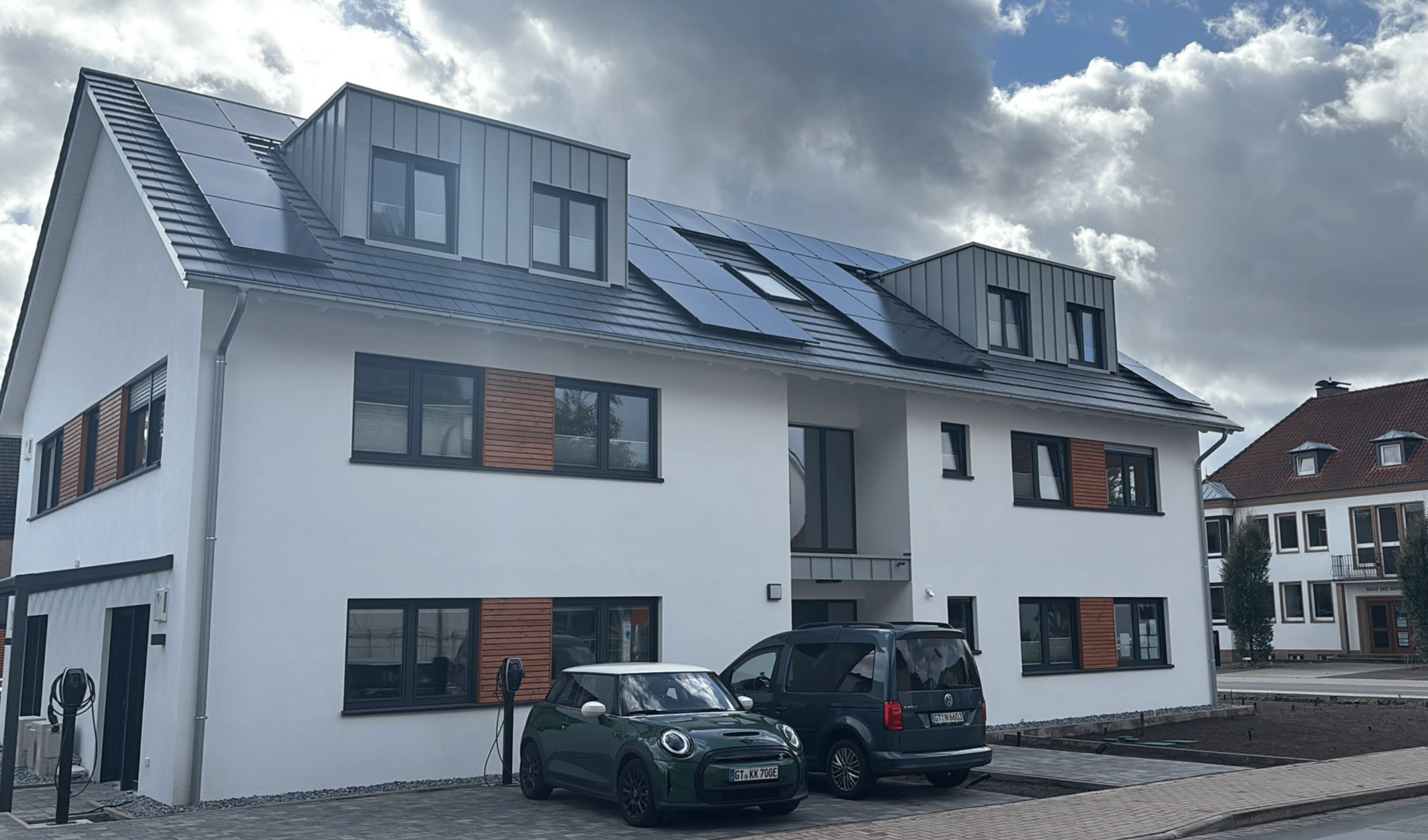 Modernes, weißes Wohnhaus mit Solarpaneelen auf dem Dach und zwei Elektroautos davor.