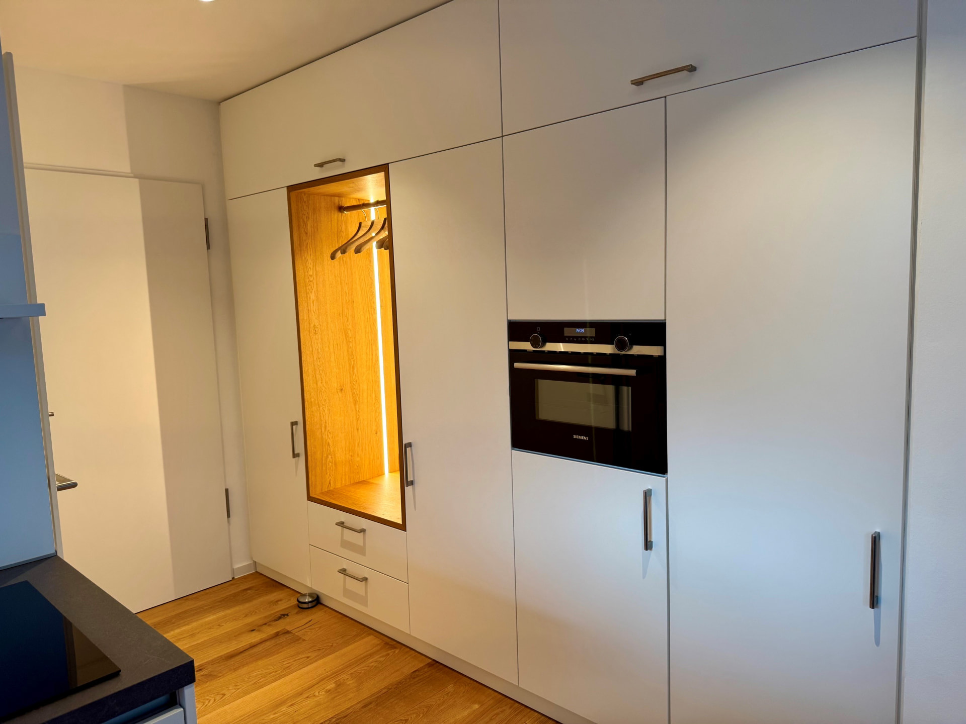 Schrankwand in Weiß mit integriertem Backofen, Kühlschrank und beleuchteter Garderobennische in Eiche.