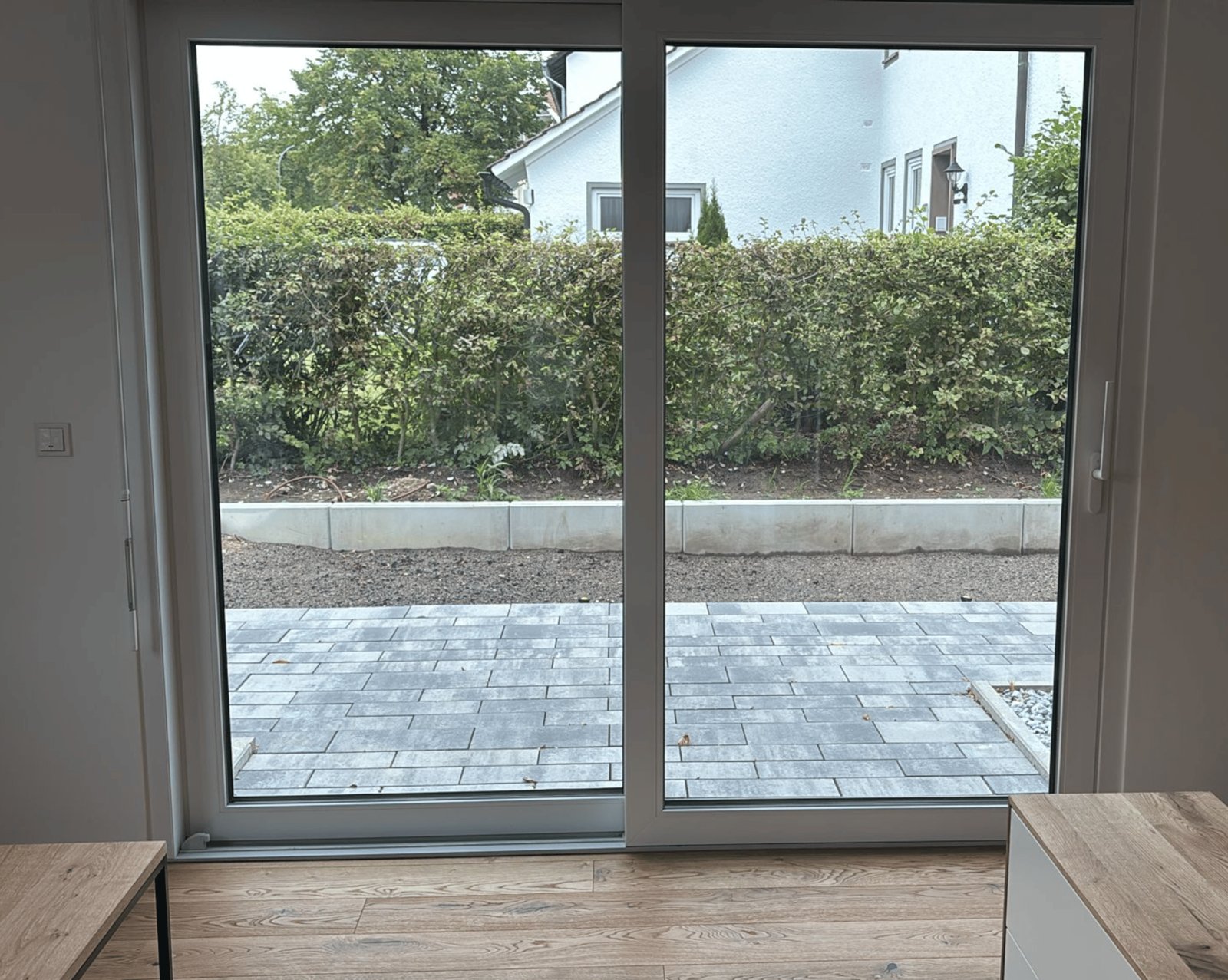 Blick durch eine Glasschiebetür auf eine gepflasterte Terrasse, Hecke und ein Haus.