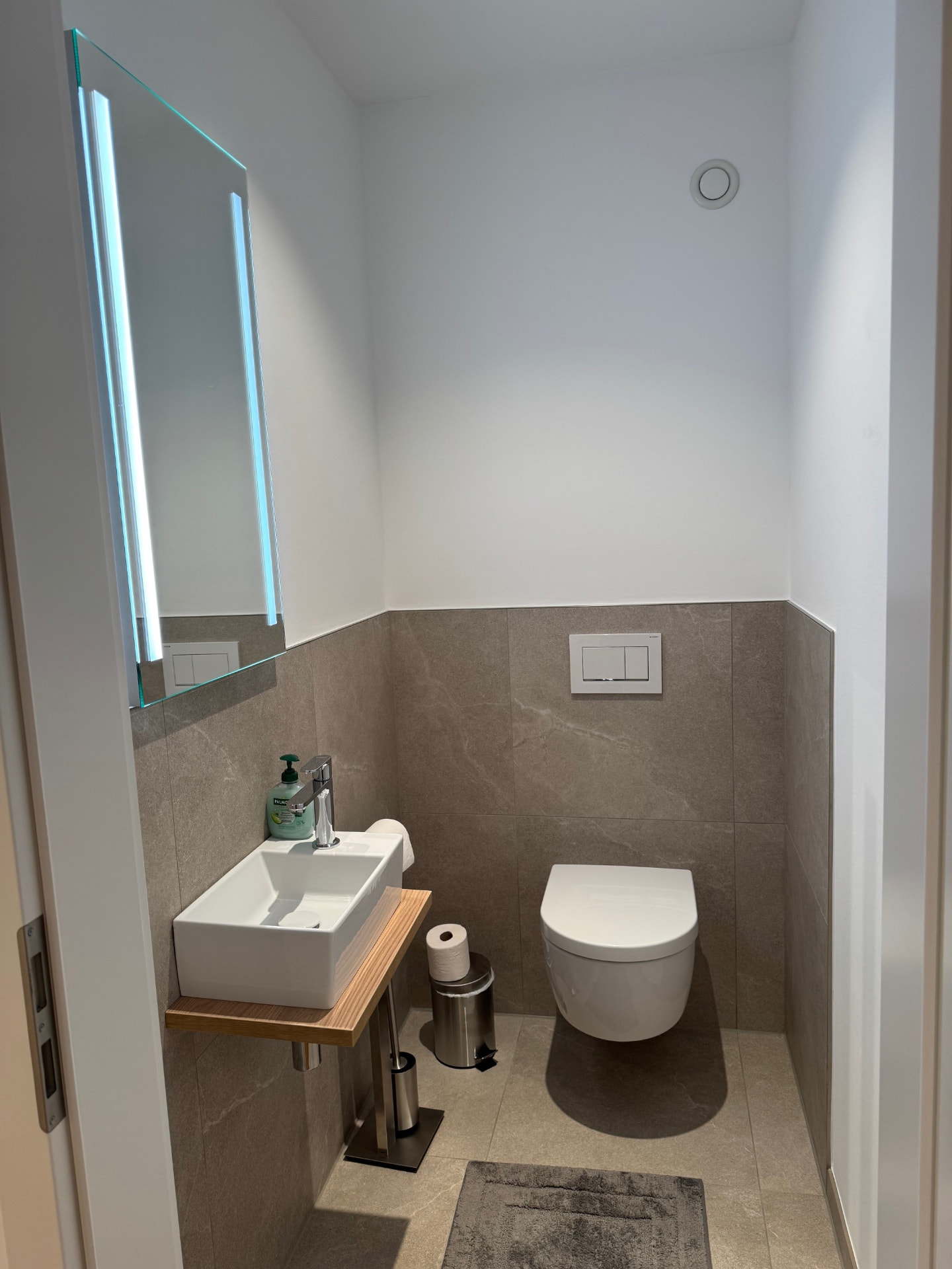 Gäste-WC mit Wand-WC, kleinem Waschbecken auf Holzkonsole und beleuchtetem Spiegel.