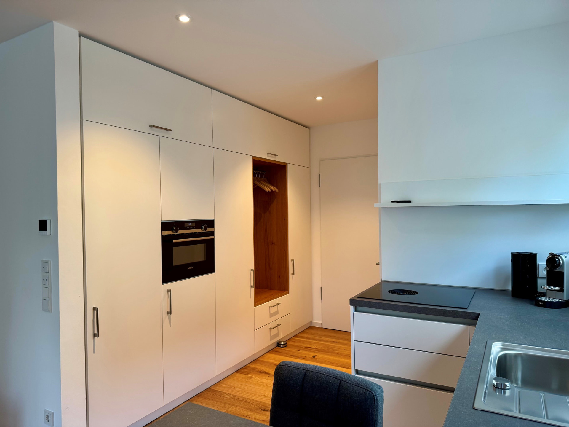 Schrankwand in Weiß mit integriertem Backofen, Kühlschrank und offener Garderobennische in Eiche.