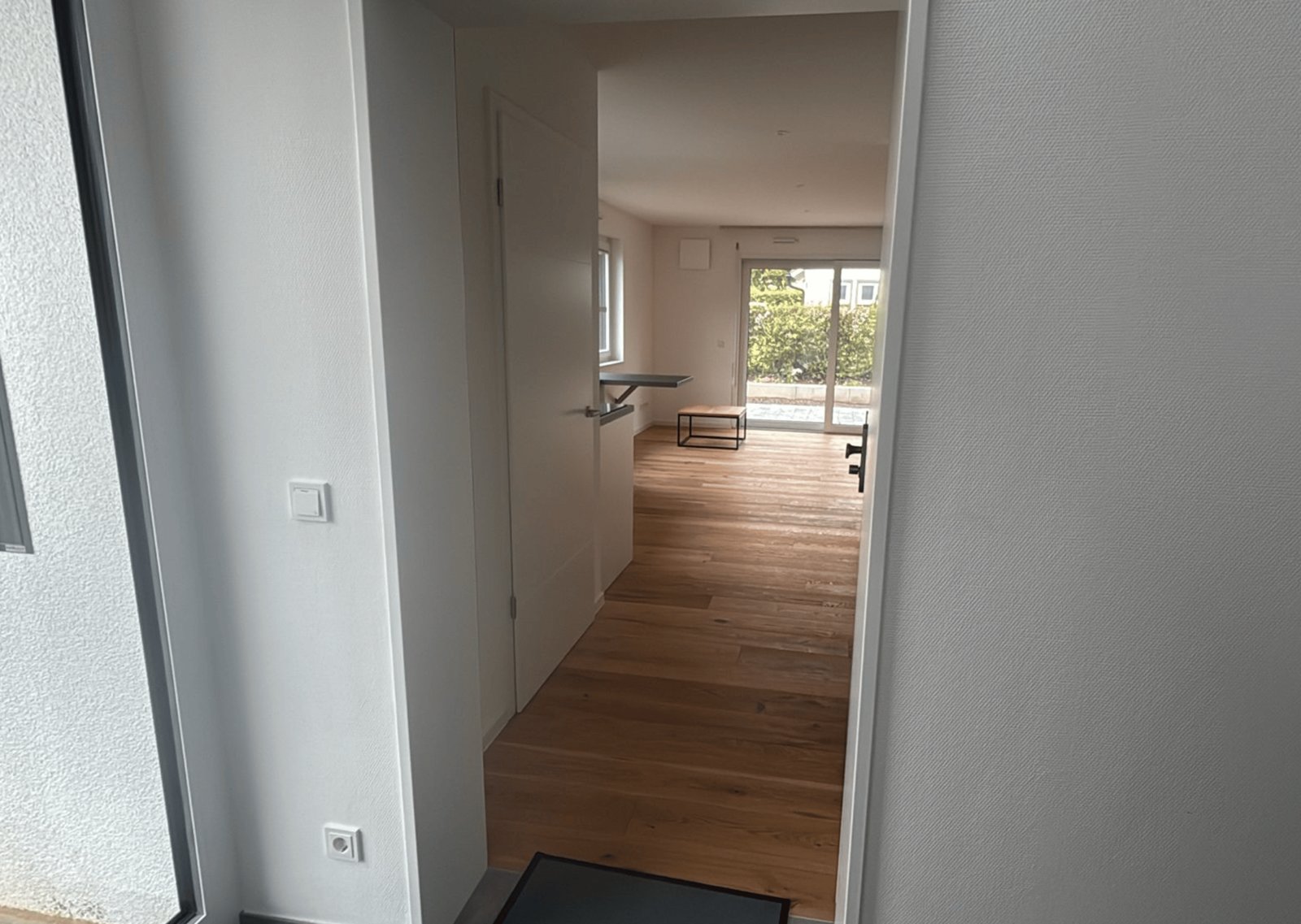 Blick durch einen Türrahmen in einen leeren, modernen Flur mit Holzboden, der zu einem Wohnzimmer führt.