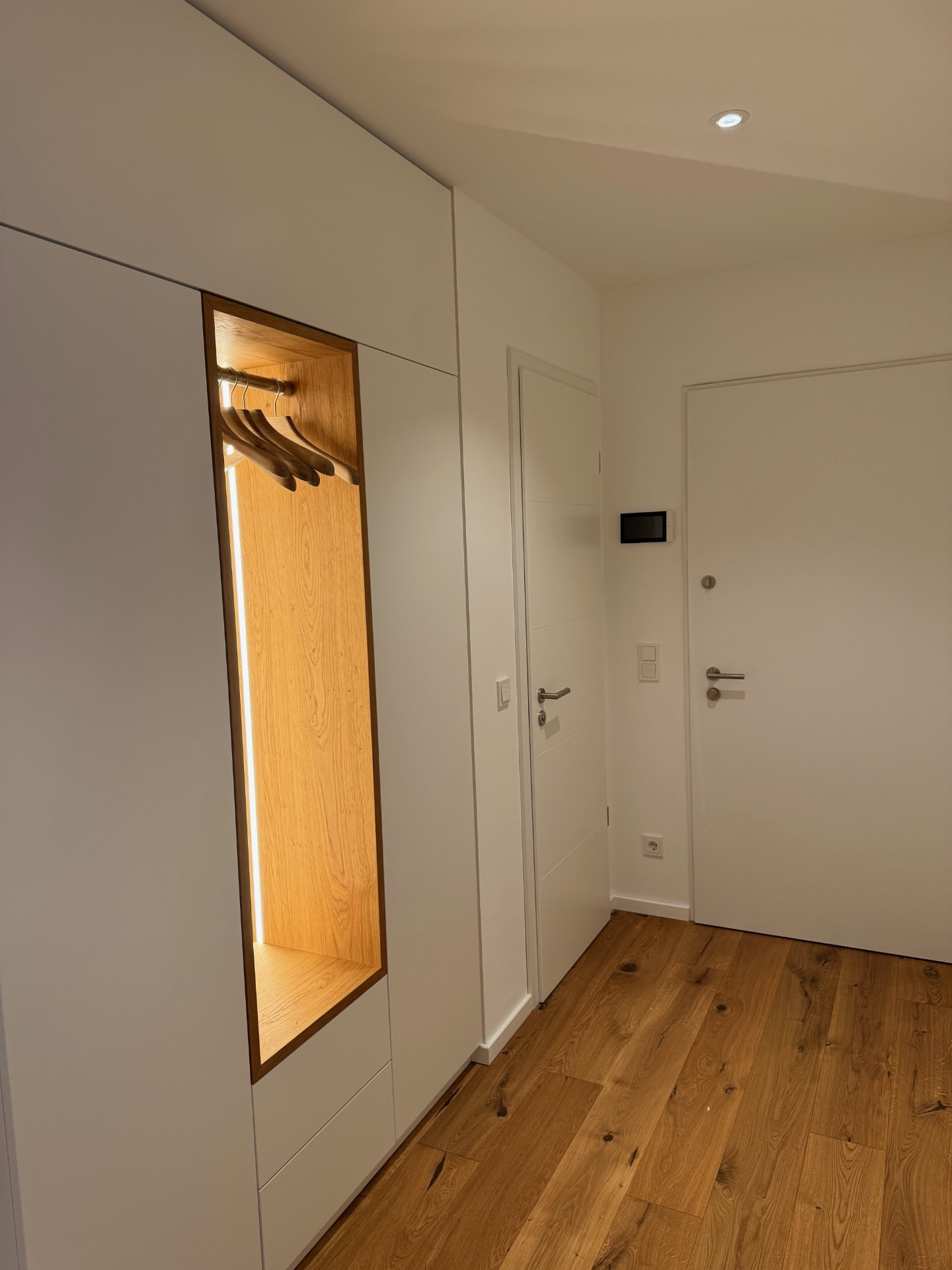 Flur mit weißer Einbau-Garderobe, beleuchteter Eichennische und Wohnungstür.