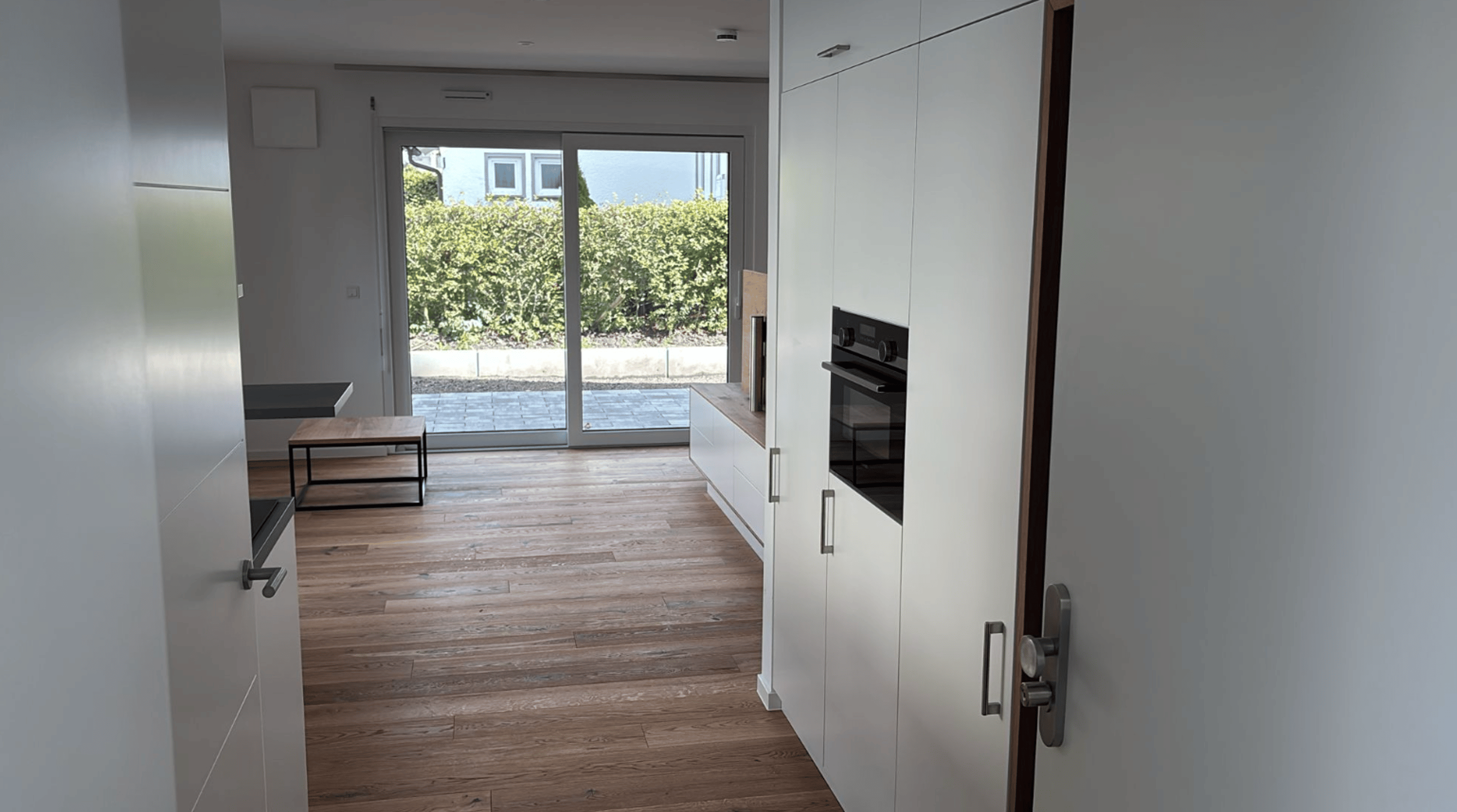 Moderne Einbauküche mit weißer Schrankfront und Blick auf Holzboden und Glastür zum Garten.