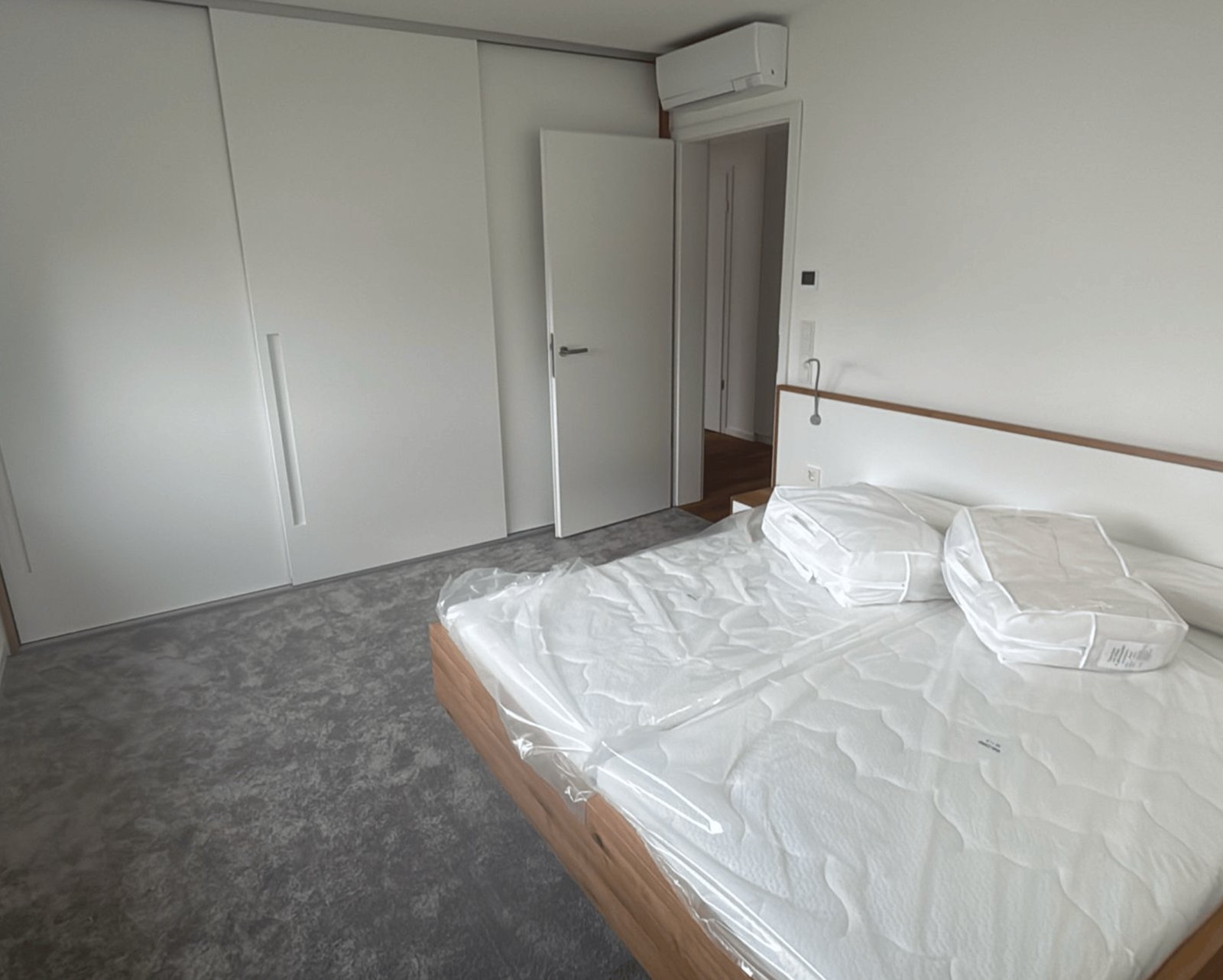 Schlafzimmer mit weißem Bett, grauem Teppich, Einbauschränken und offener Tür.