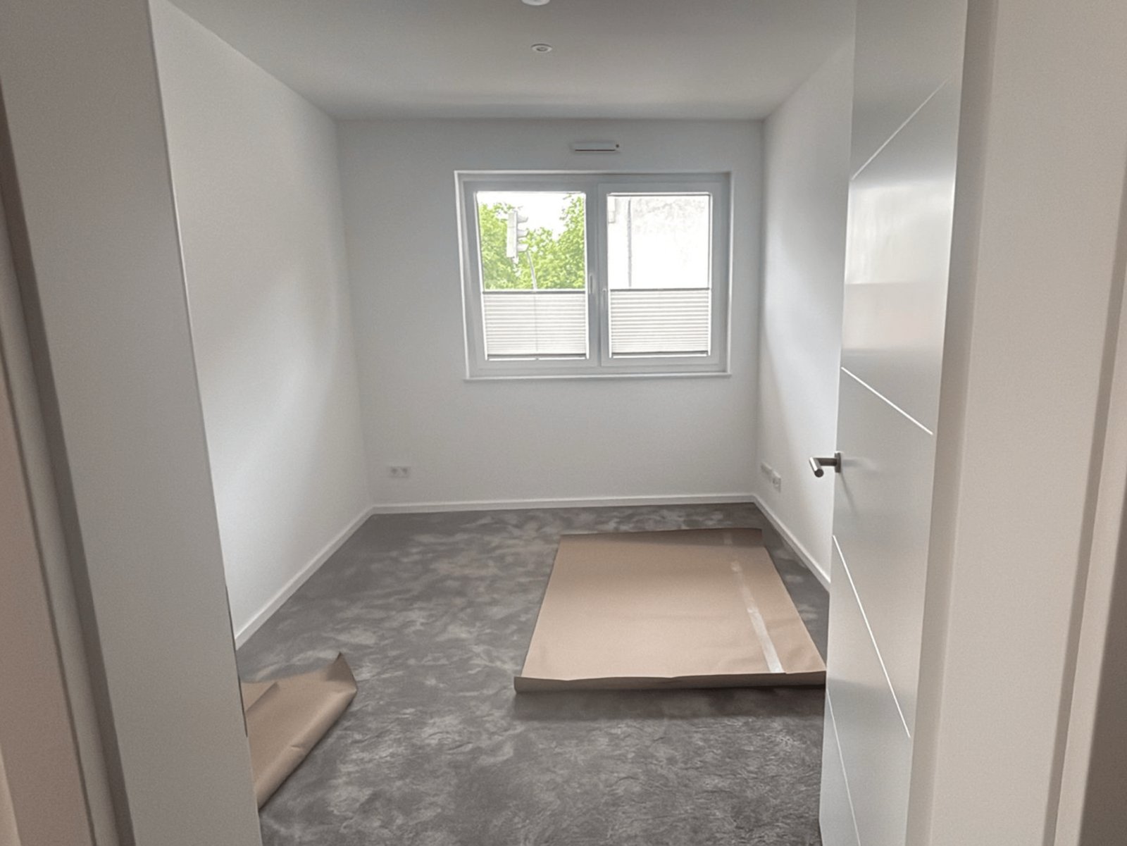 Leeres Zimmer mit grauem Teppichboden, weiß gestrichenen Wänden und einem Fenster.