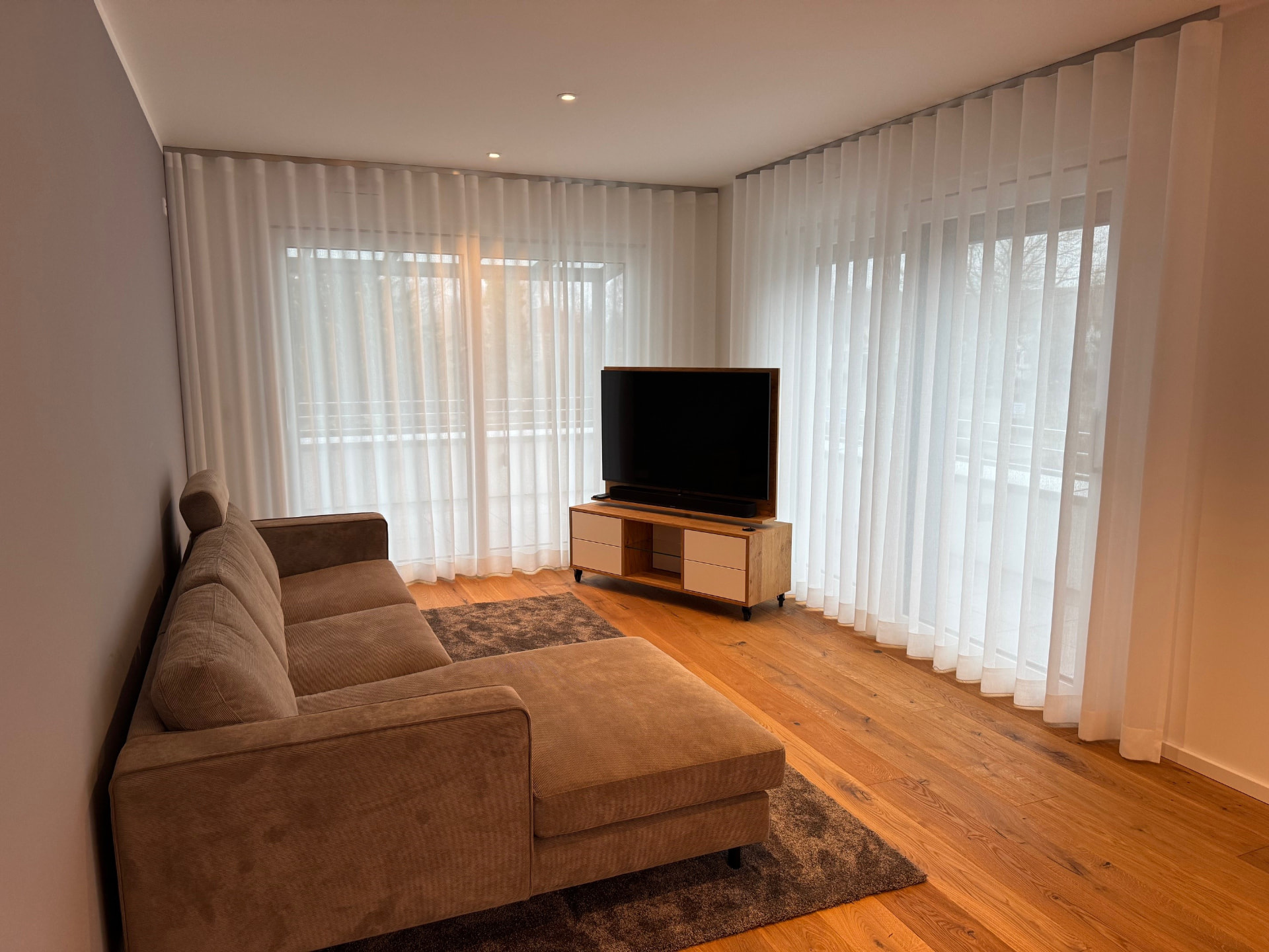 Wohnzimmer mit Cordsofa, Eichen-TV-Board und raumhohen Vorhängen an zwei Fensterfronten.