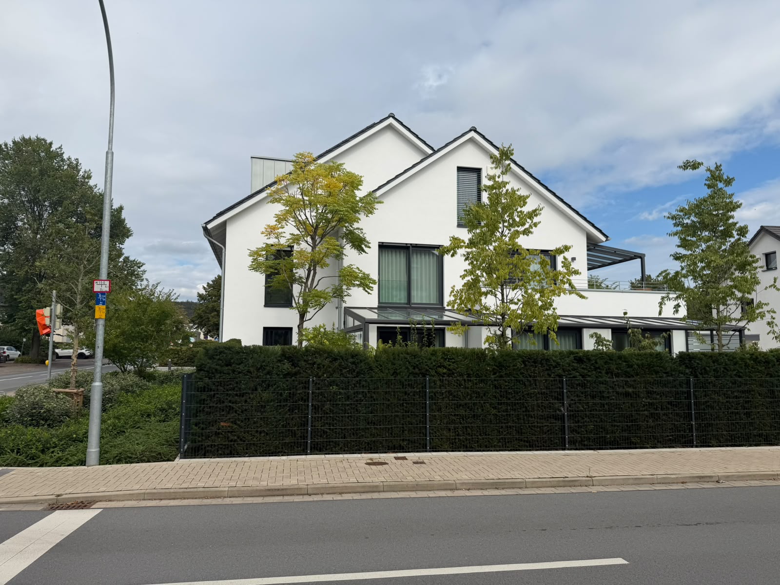 Doppelgiebel-Fassade in Weiß mit gläsernem Terrassendach und grüner Hecke an der Straße.