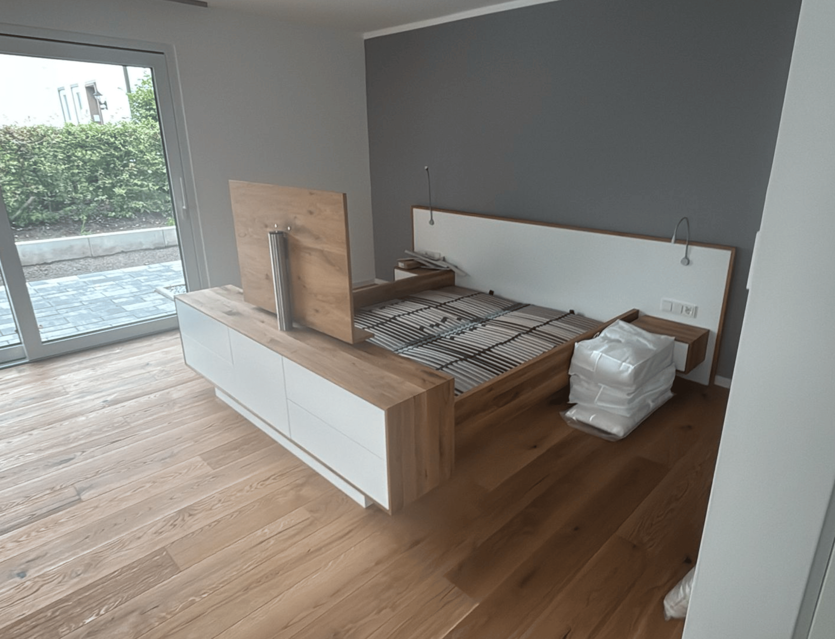 Schlafzimmer mit Holzboden, Bett mit weißem Kopfteil und Sideboard mit ausfahrbarem Fernseher.