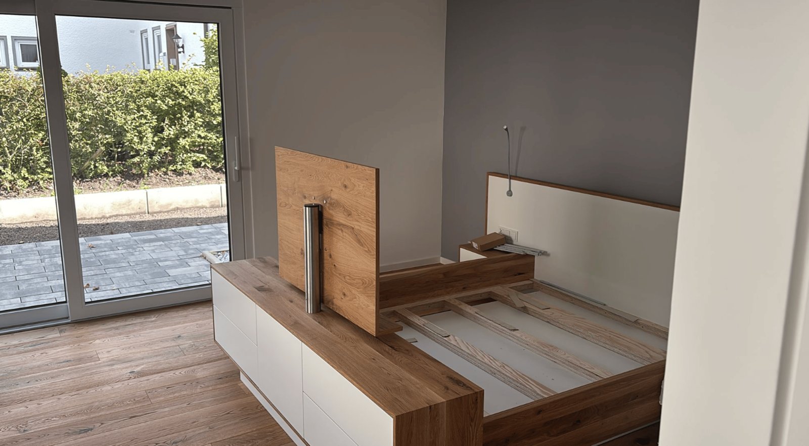 Holzbett mit weißem Kopfteil und Schubladen in einem leeren Raum mit Glastür.